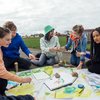 Gruppe von Jugendlichen sitzt im Freien und bemalt und besprayt ein buntes Tuch am Boden