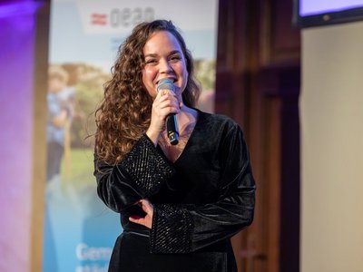 Junge Frau mit langen Haaren und schwarzem Oberteil spricht mit Mikrophon in der Hand
