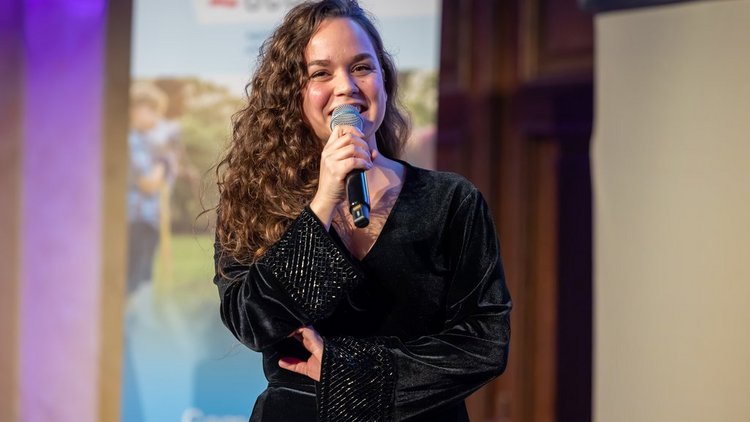 Junge Frau mit langen Haaren und schwarzem Oberteil spricht mit Mikrophon in der Hand
