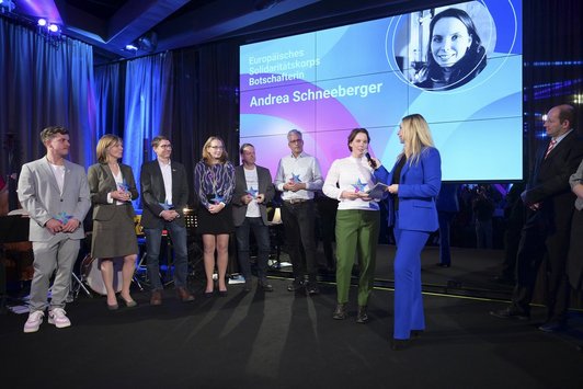 Neun Personen stehen auf der Bühne, eine Dame hält einer anderen Dame das Mikrofon hin. Im Hintergrund am Screen der Schriftzug "Europäisches Solidaritätskorps Botschafterin Andrea Schneeberger"