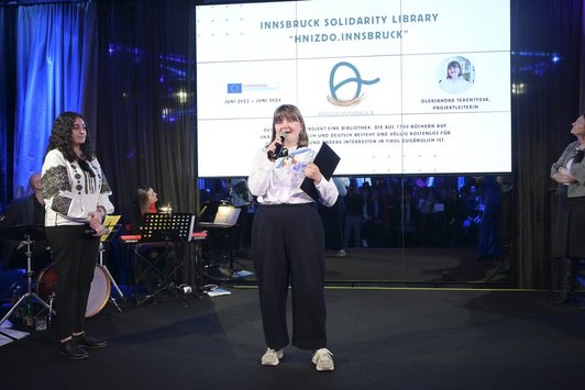 Eine Frau steht auf der Bühne und spricht in ein Mikrofon, dahinter ein Screen mit dem Schriftzug „Innsbruck Solidarity Library“