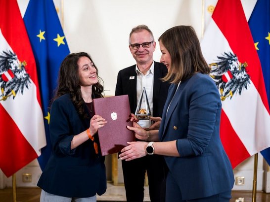 Ministerin überreicht junger Frau eine rote Urkundenmappe mit Wappen im Hintergrund Mann mit Brille