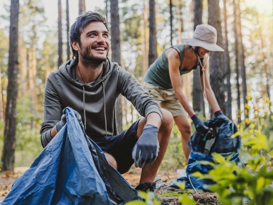 Junger Mann mit blauer Mülltüte im Wald, hinter im andere Person ebenfalls am Müllsammeln mit Hut